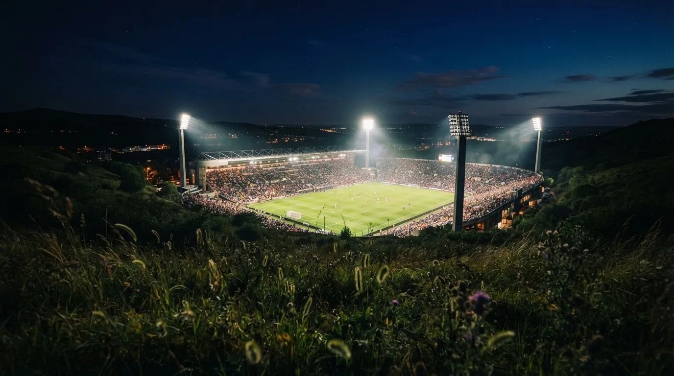 Ein Stadion unter Flutlicht bei Nacht, von einem Hügel aus der Ferne betrachtet, mit Dunst über dem Spielfeld.