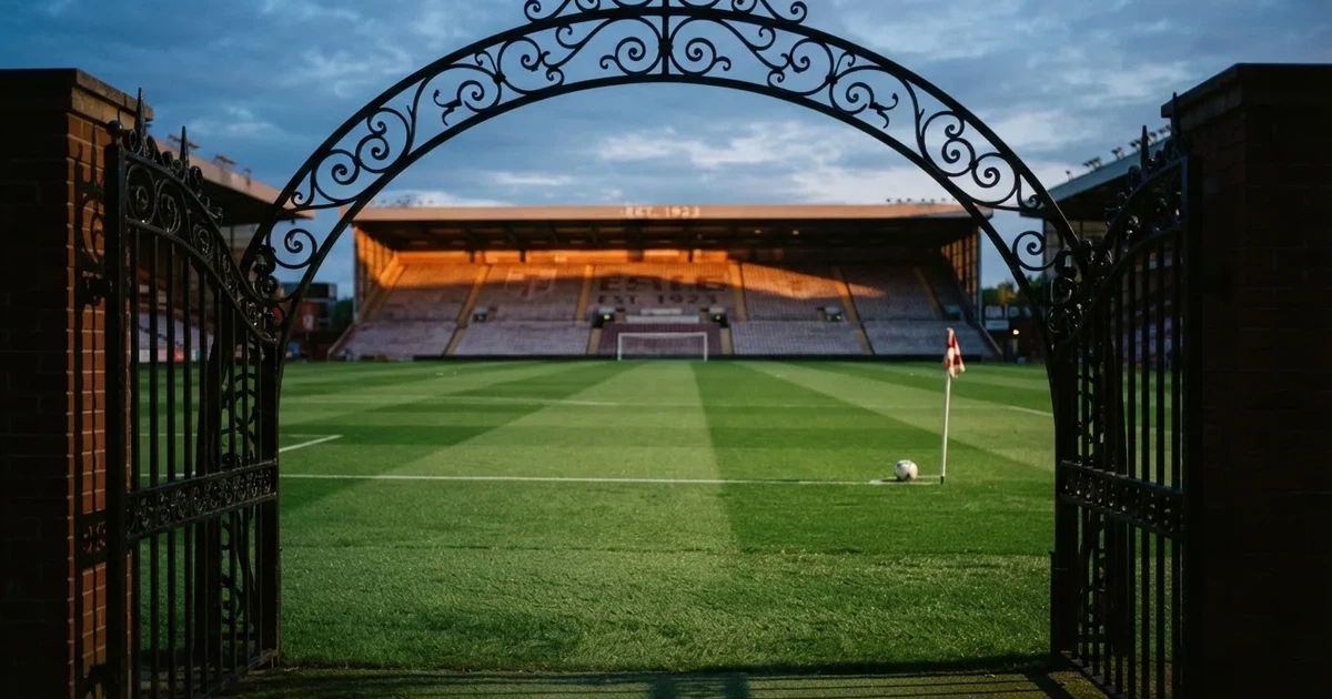 Blick durch ein Stadiontor auf einen grünen Rasen im Abendlicht, ein Ball liegt an der Eckfahne, die Ränge sind leer.