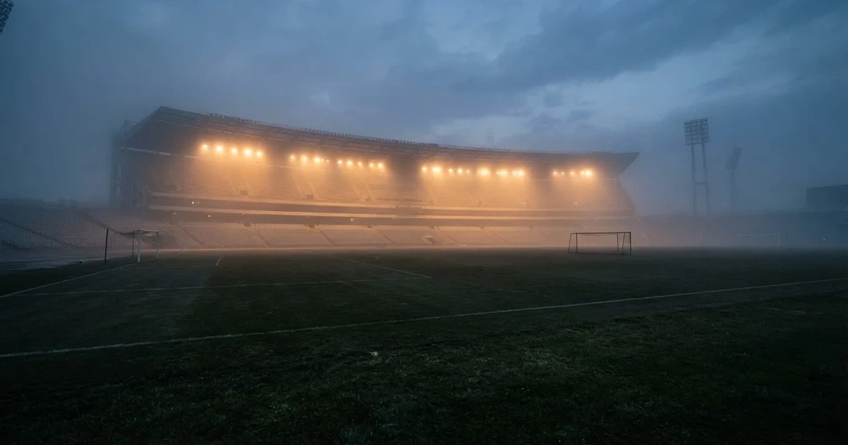 Ein Stadion unter Flutlicht, aus großer Entfernung über eine dunkle Wiese fotografiert, am Himmel zieht Nebel auf.